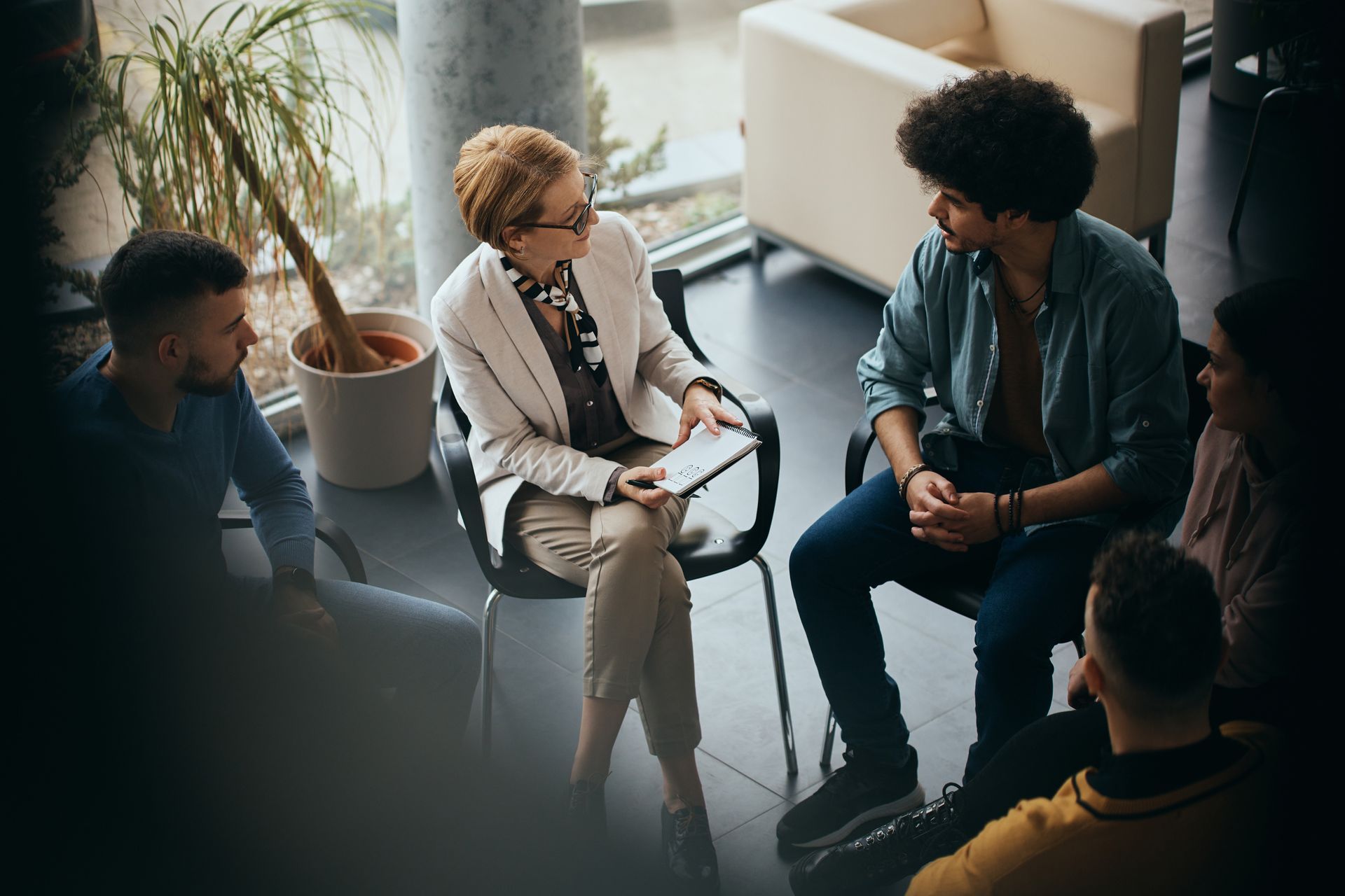 Small group in a circle meeting, led by a woman in a white suit in a bright office lounge.