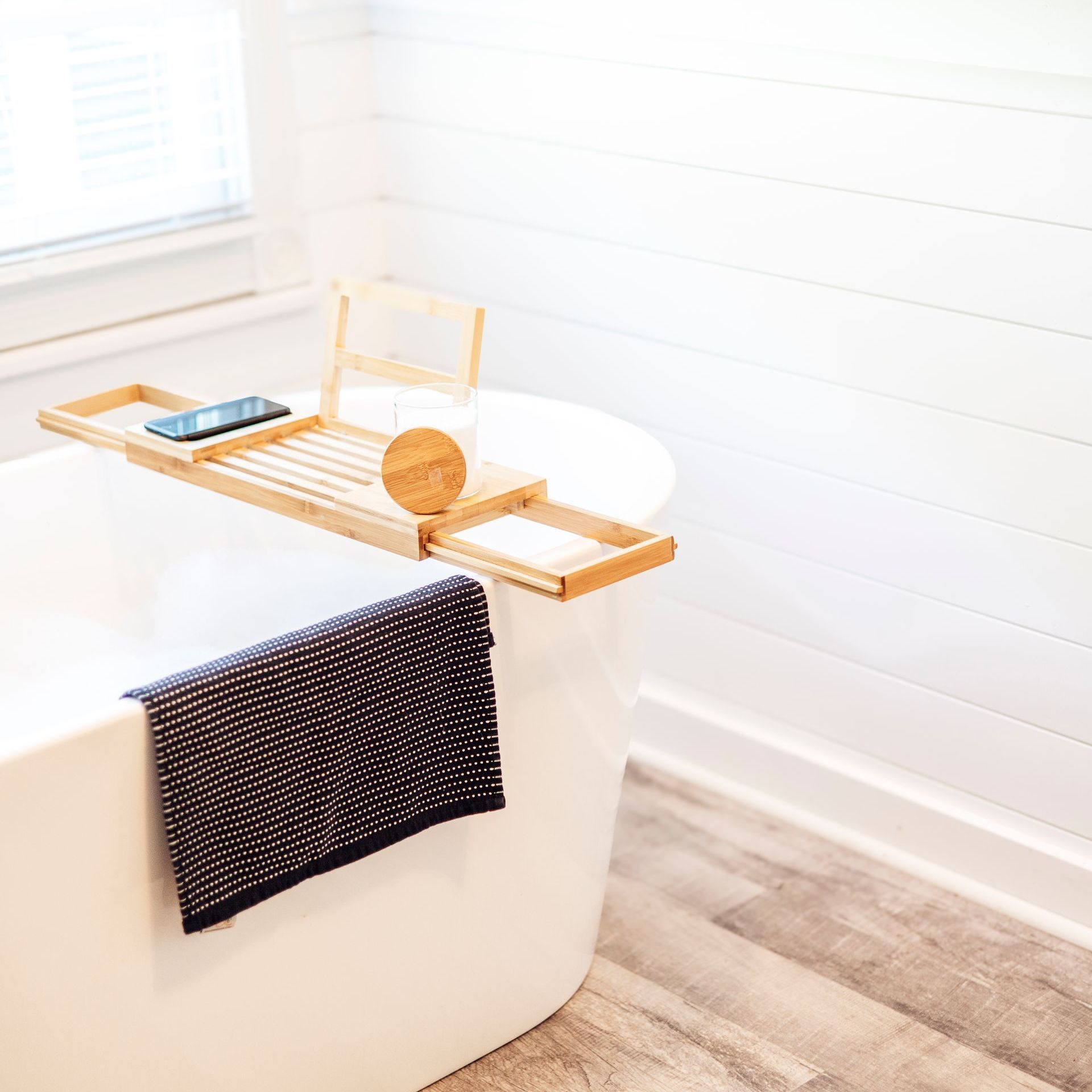 A white bathtub with a wooden tray holding a phone, candle, and other items. A dark towel hangs on the side.