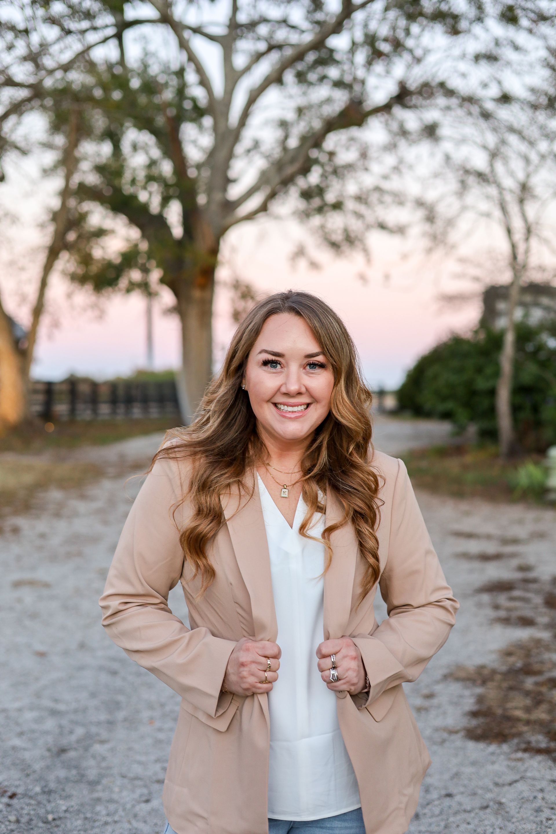 Woman smiling, wearing beige blazer, white top, standing outdoors in front of trees.