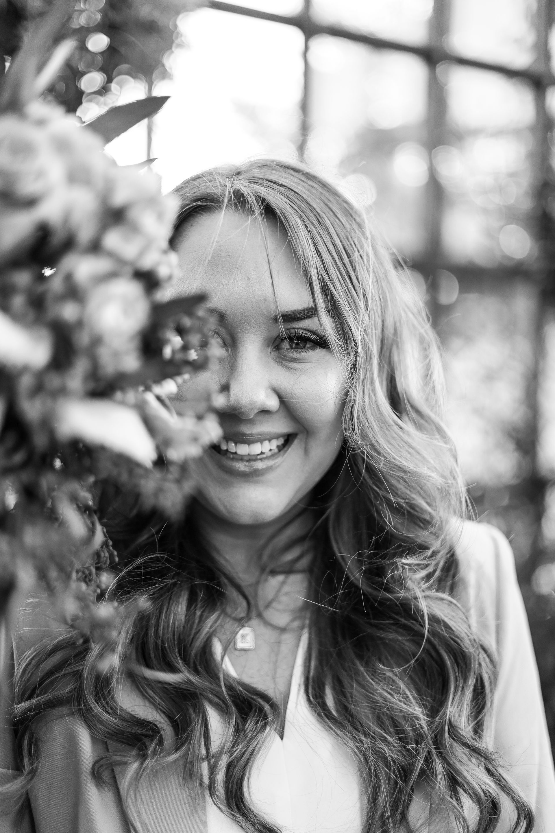Woman smiling, partially obscured by flowers. Long wavy hair, jacket, bright background.