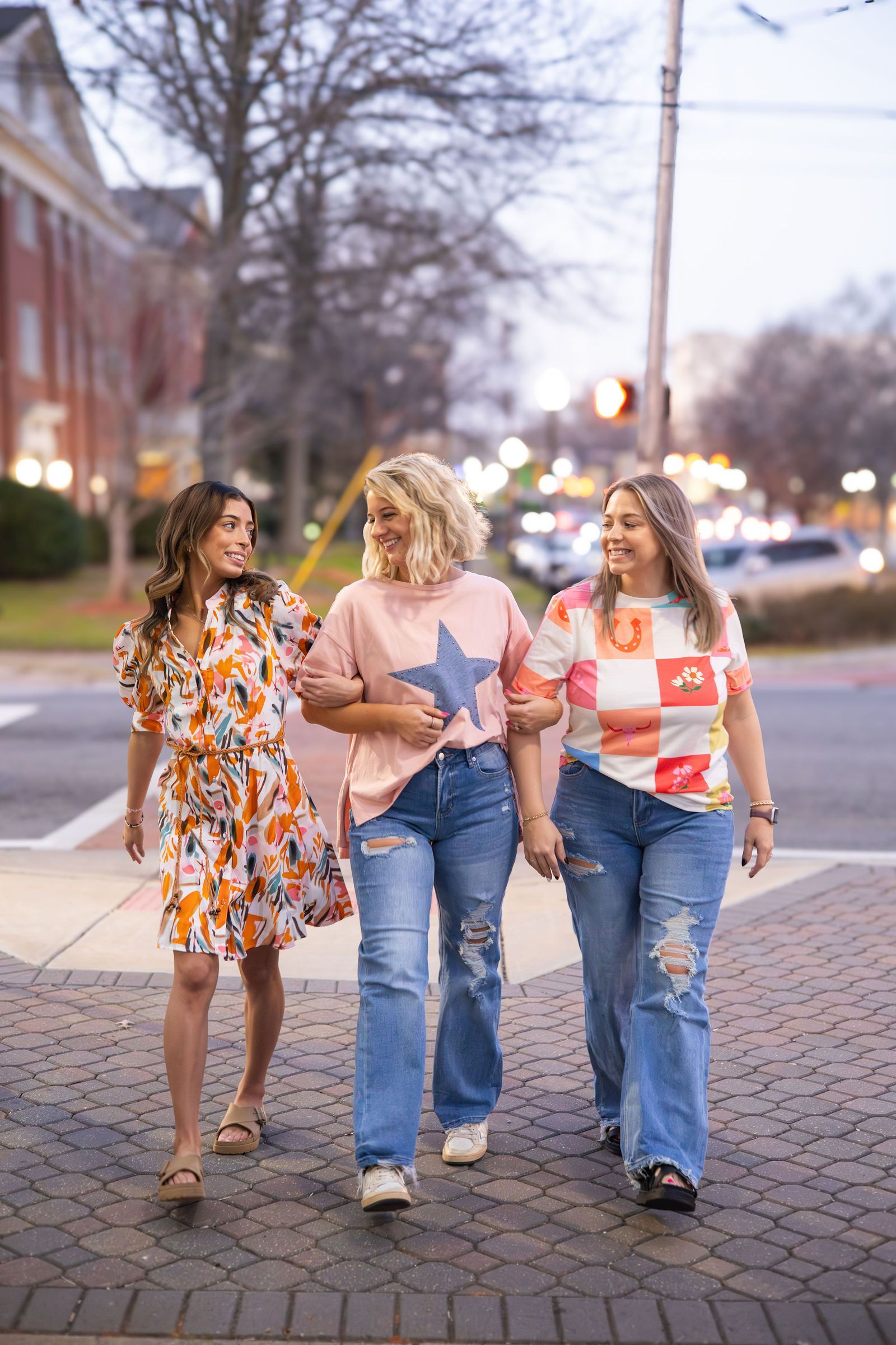 Three people arm in arm, crossing a street. One wears a floral dress, another a pink star shirt and distressed jeans, and the last a colorful block shirt and jeans.