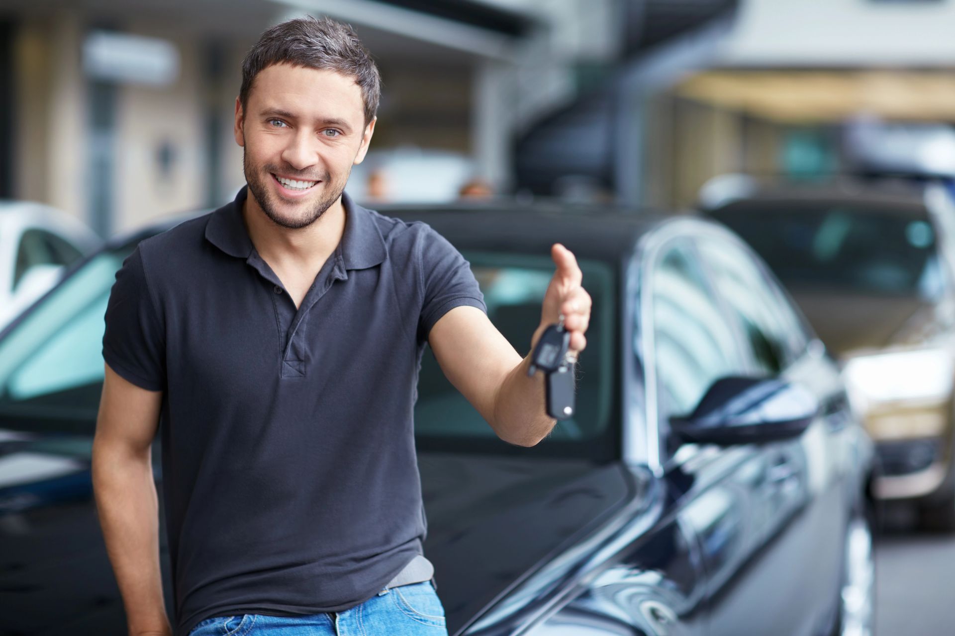 Smiling car salesman holding keys beside a black sedan in a dealership lot