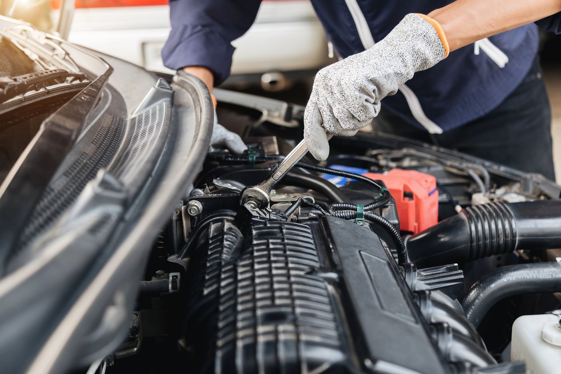 Mechanic using pliers to repair wiring in a car engine bay, with a red battery visible