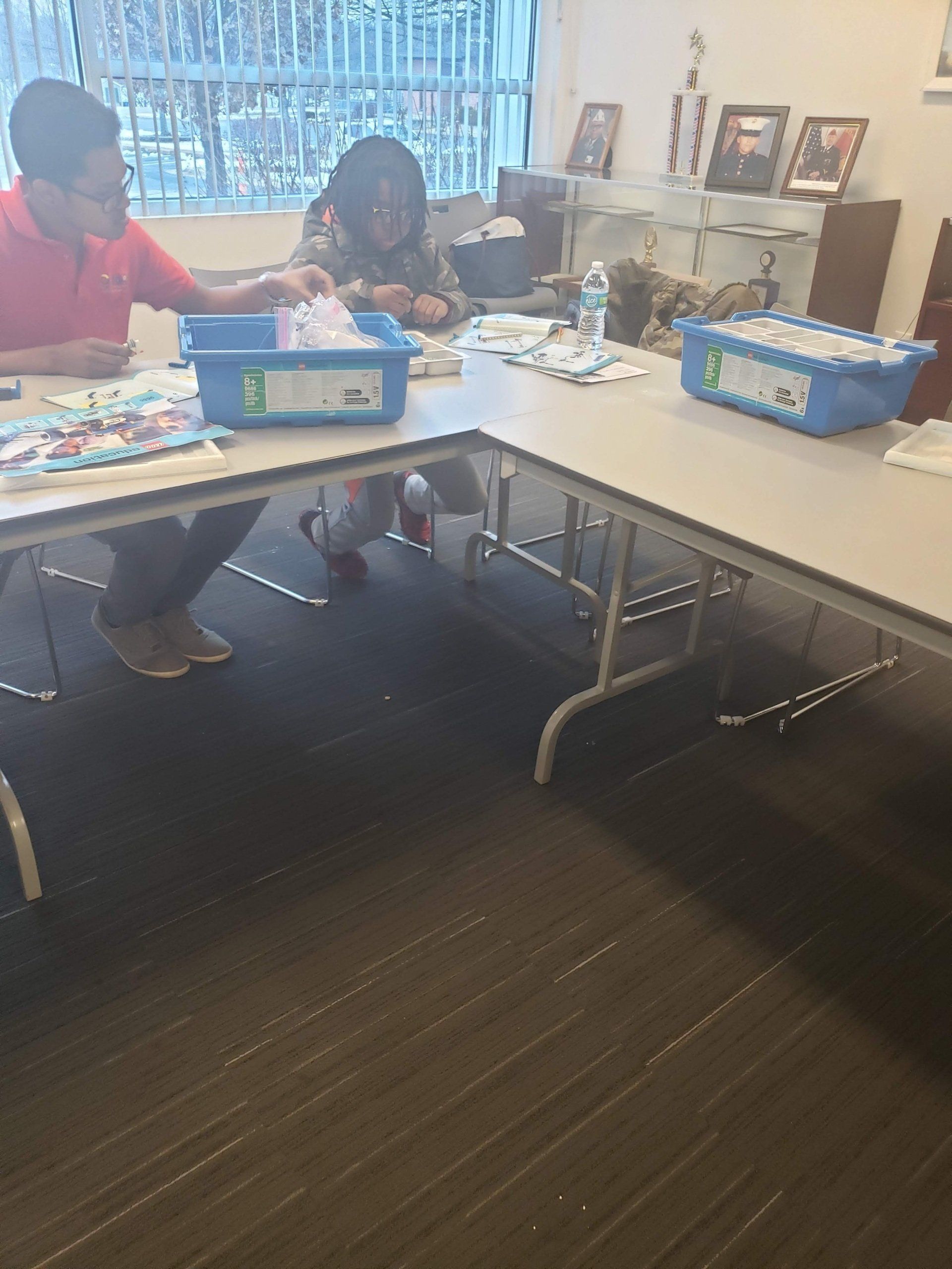 Two people sit at a corner table in an office, sorting through items into blue plastic bins.
