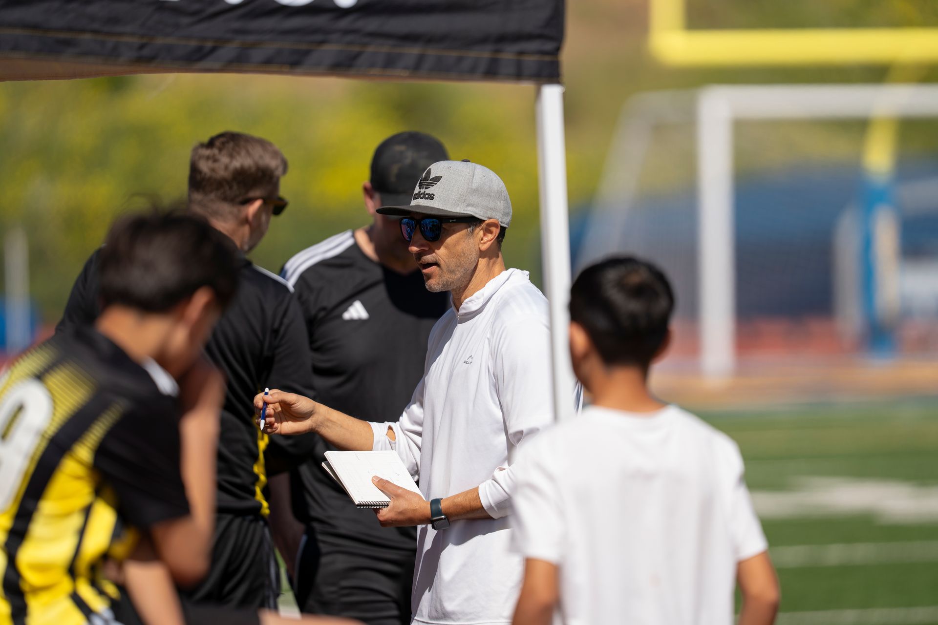 Soccer coach talking to players on a sunny field, holding a clipboard.