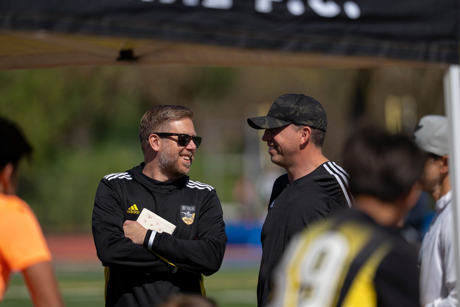 Two men in black tracksuits converse on a soccer field; one wears sunglasses and holds a notepad.