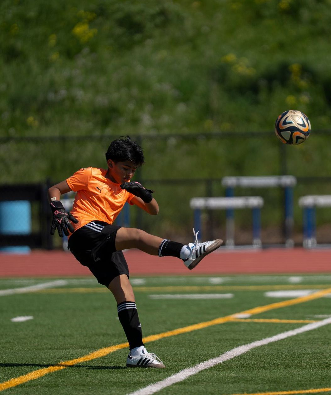 Soccer player in orange shirt kicking ball on a green field. Black shorts, white shoes. Goal in the background.