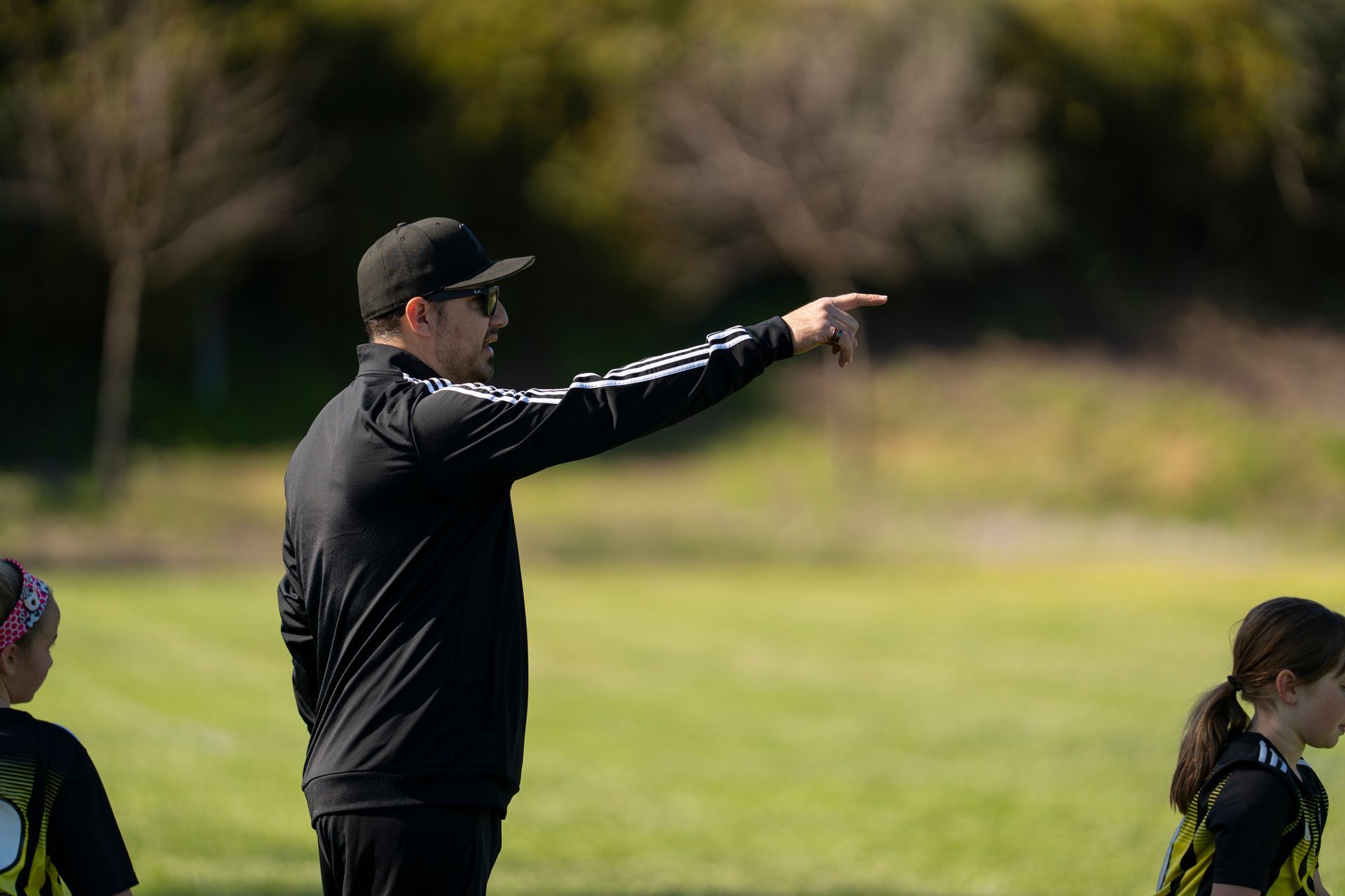 Soccer coach in black jacket and cap points to the right on a green field.