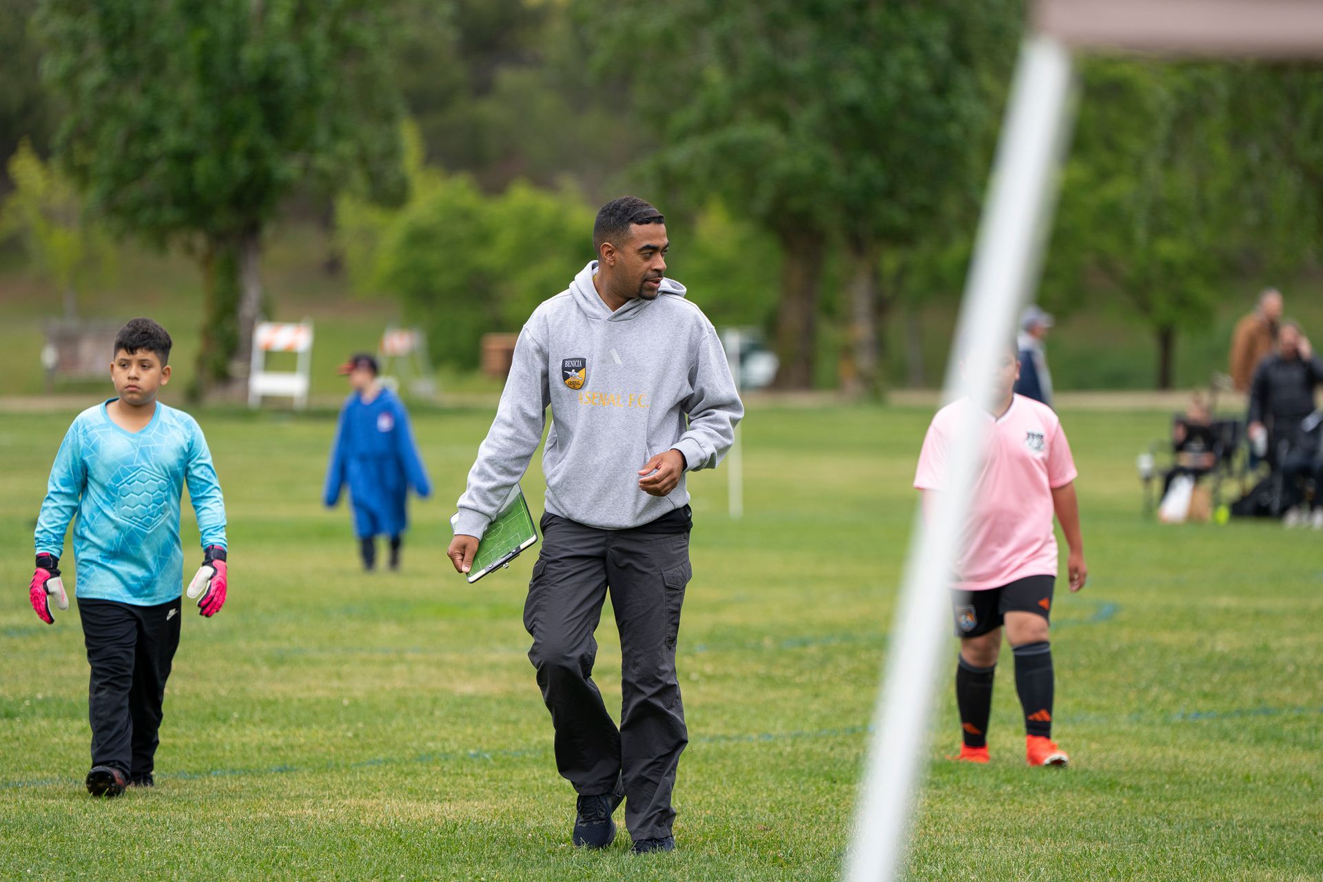 Soccer coach walking on a field with two young players. The coach holds a clipboard, and players are in uniform.