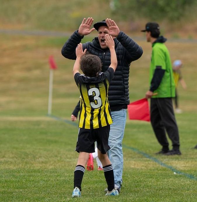 Soccer player in yellow and black uniform high-fives a man on a field; another person watches nearby.