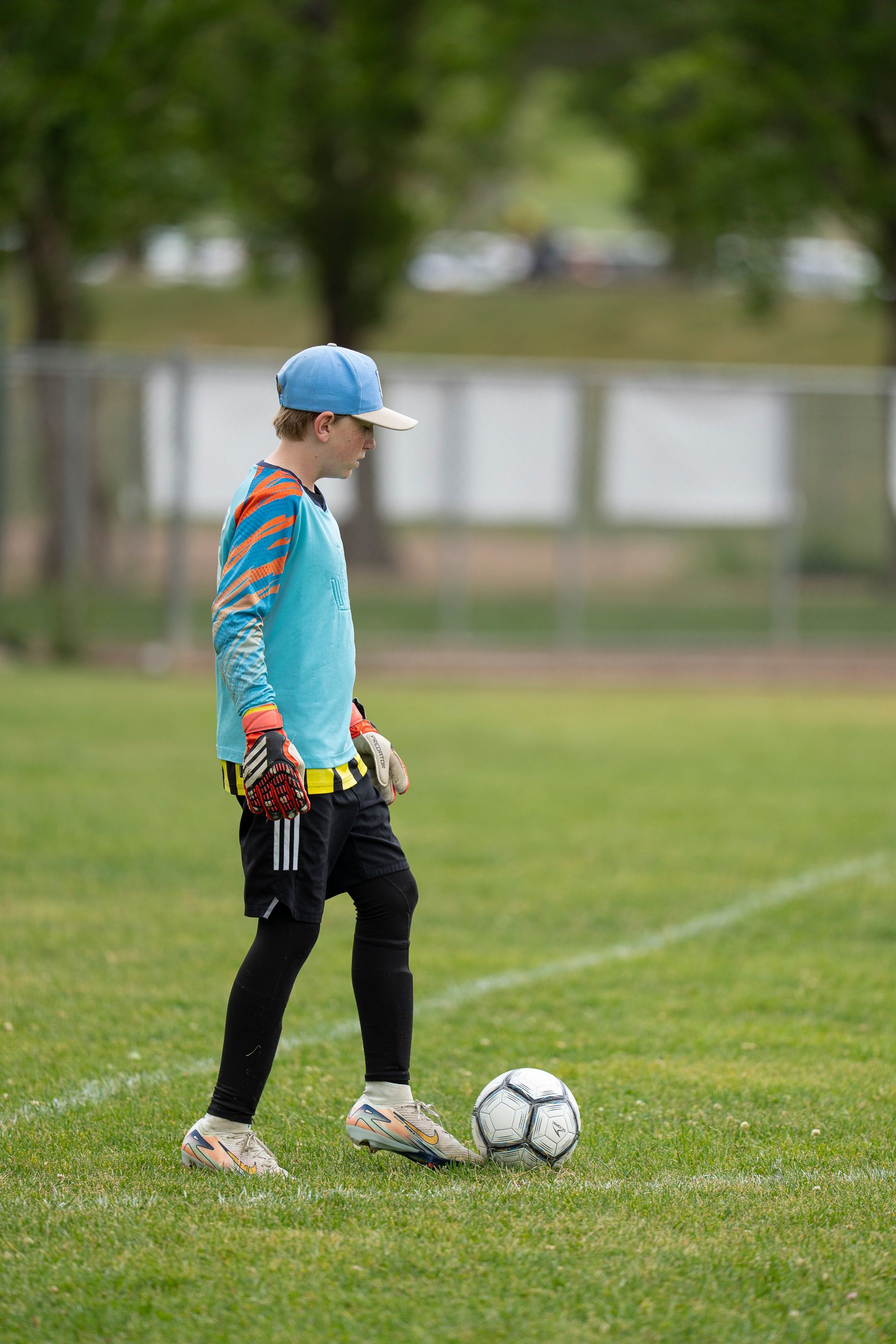 Soccer goalie in blue and orange jersey, black leggings, and blue cap, stands near the ball on a grass field.