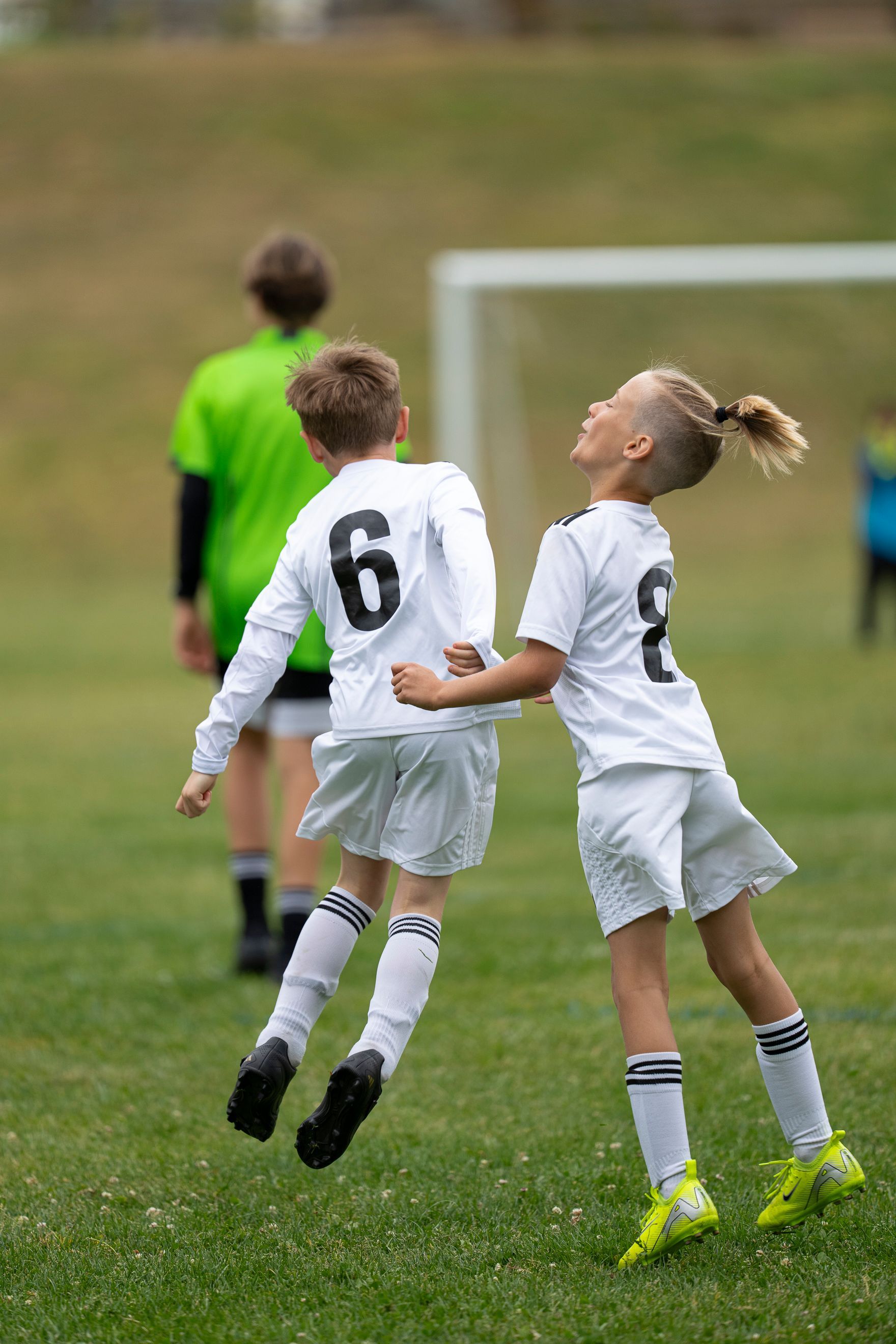 Two soccer players, wearing white jerseys, jumping in the air on a green field. A goal and another player are in the background.
