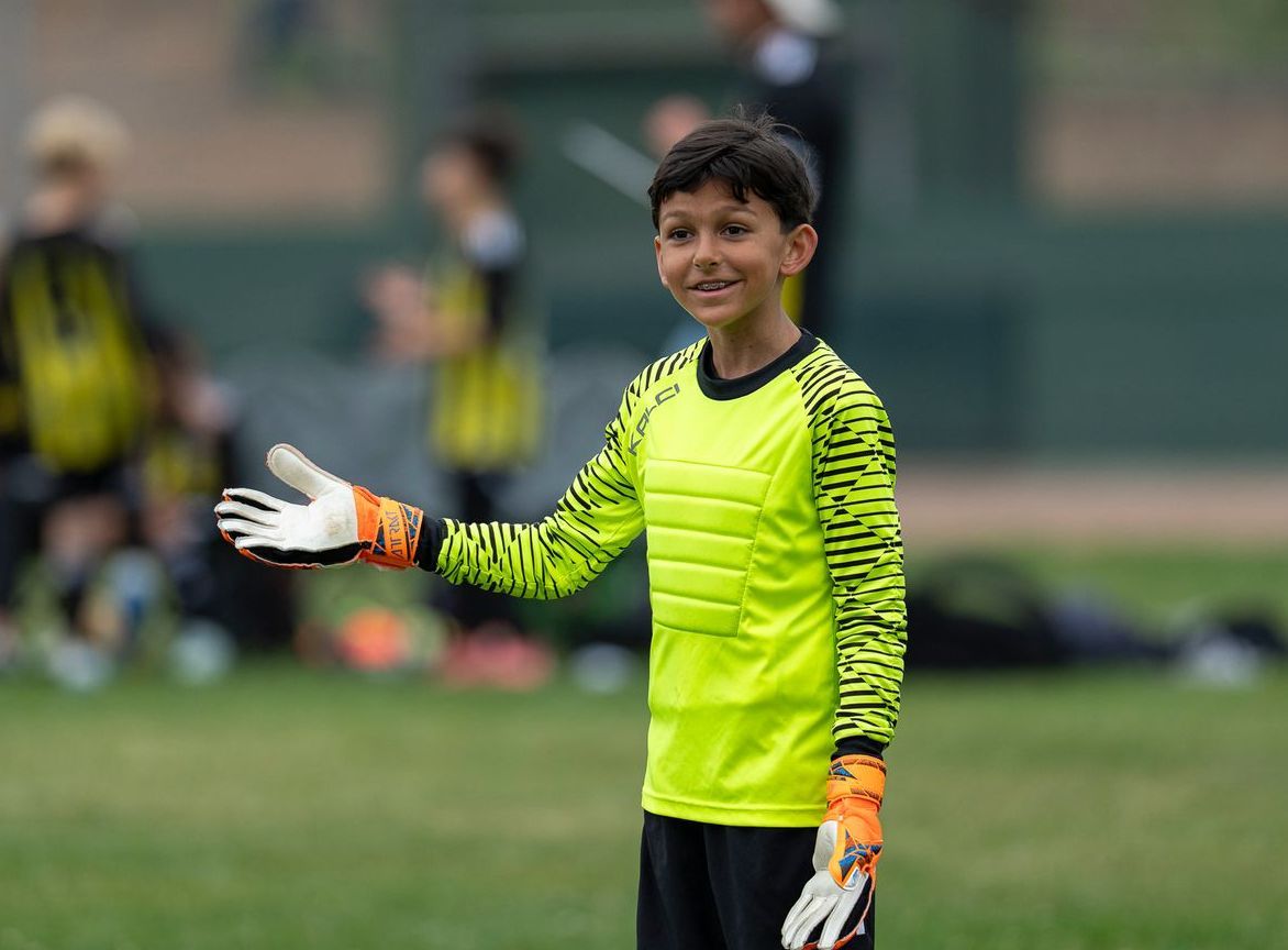 Boy soccer goalie in neon yellow jersey with orange gloves, smiling on a field.