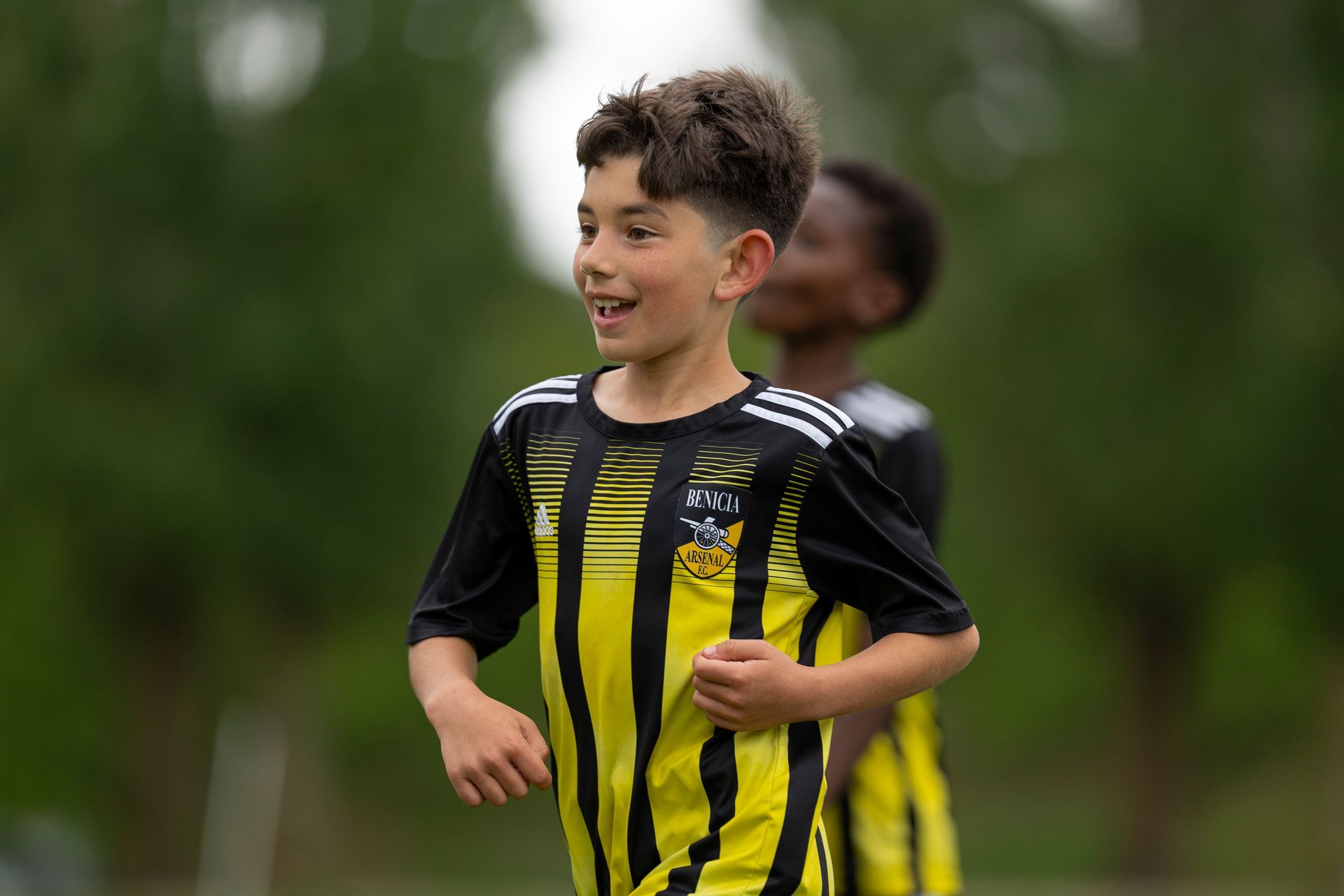 Boy in yellow and black soccer jersey smiles on a field, another person in the background.