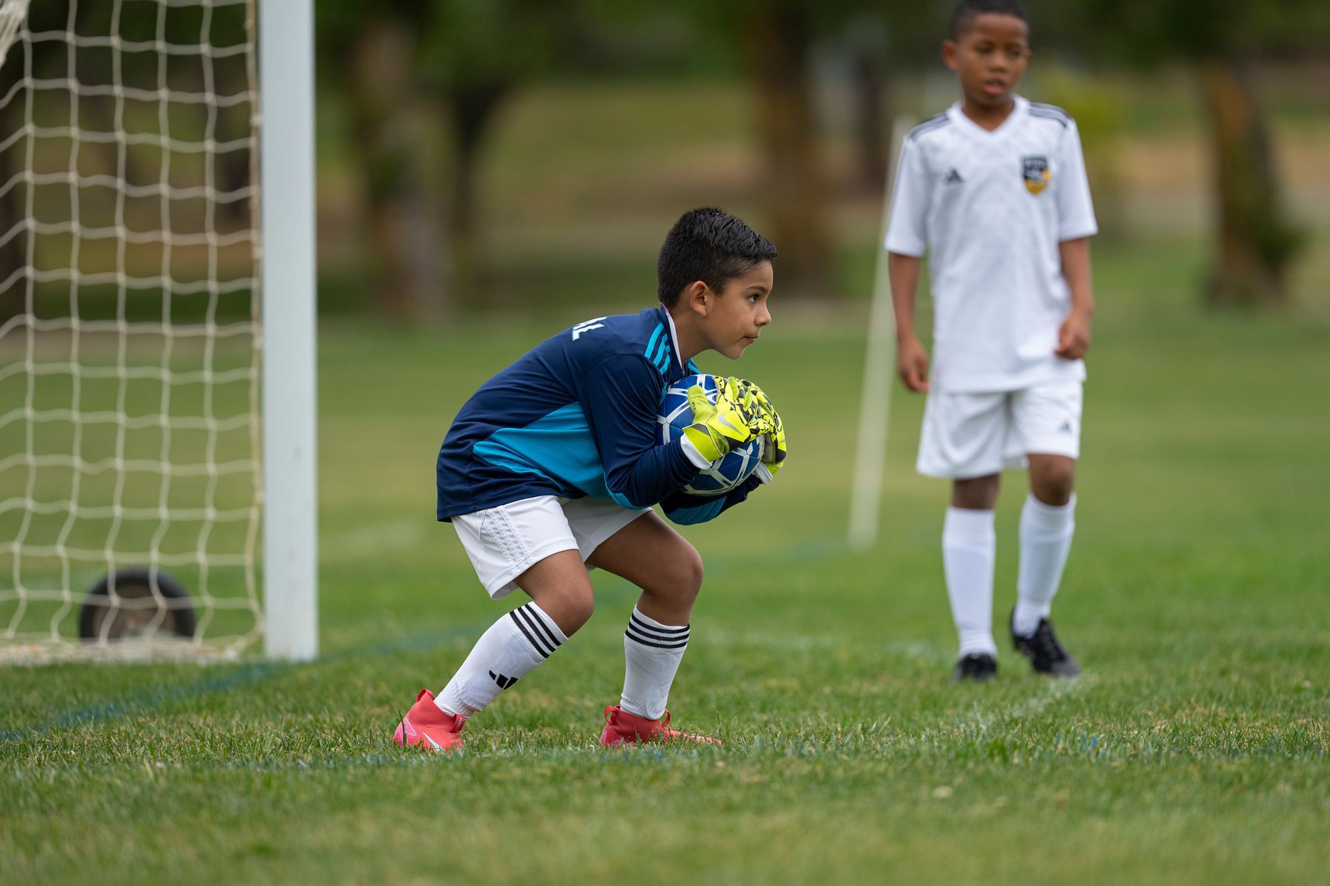 Boy soccer goalie in blue jersey, ready to catch ball in front of goal. Another player stands behind.