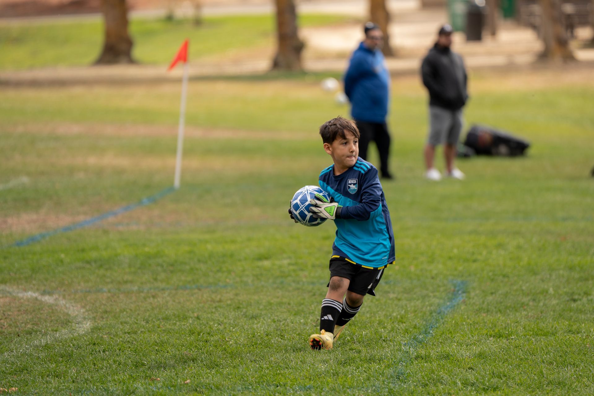 Boy in teal soccer uniform, holding ball, running on grass field. Two adults stand in the background.