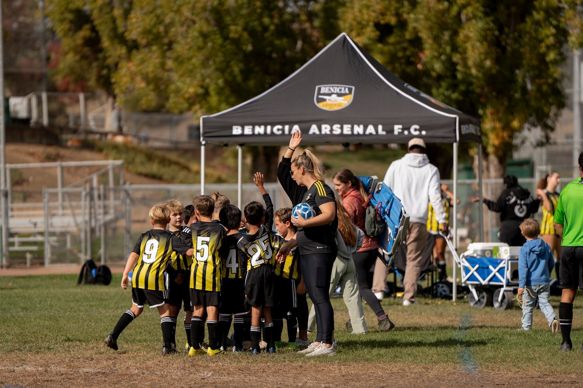Youth soccer team in black and gold uniforms huddling with coach on a grassy field, under Benicia Arsenal F.C. tent.