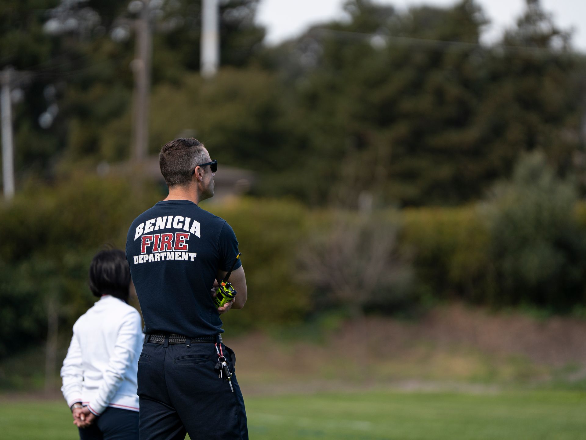 Man in a Genicia Fire Department shirt watches, a child beside him, on a grassy field with trees behind.