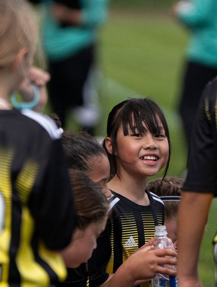 Girl in soccer uniform smiles, holding water bottle, surrounded by teammates on a field.