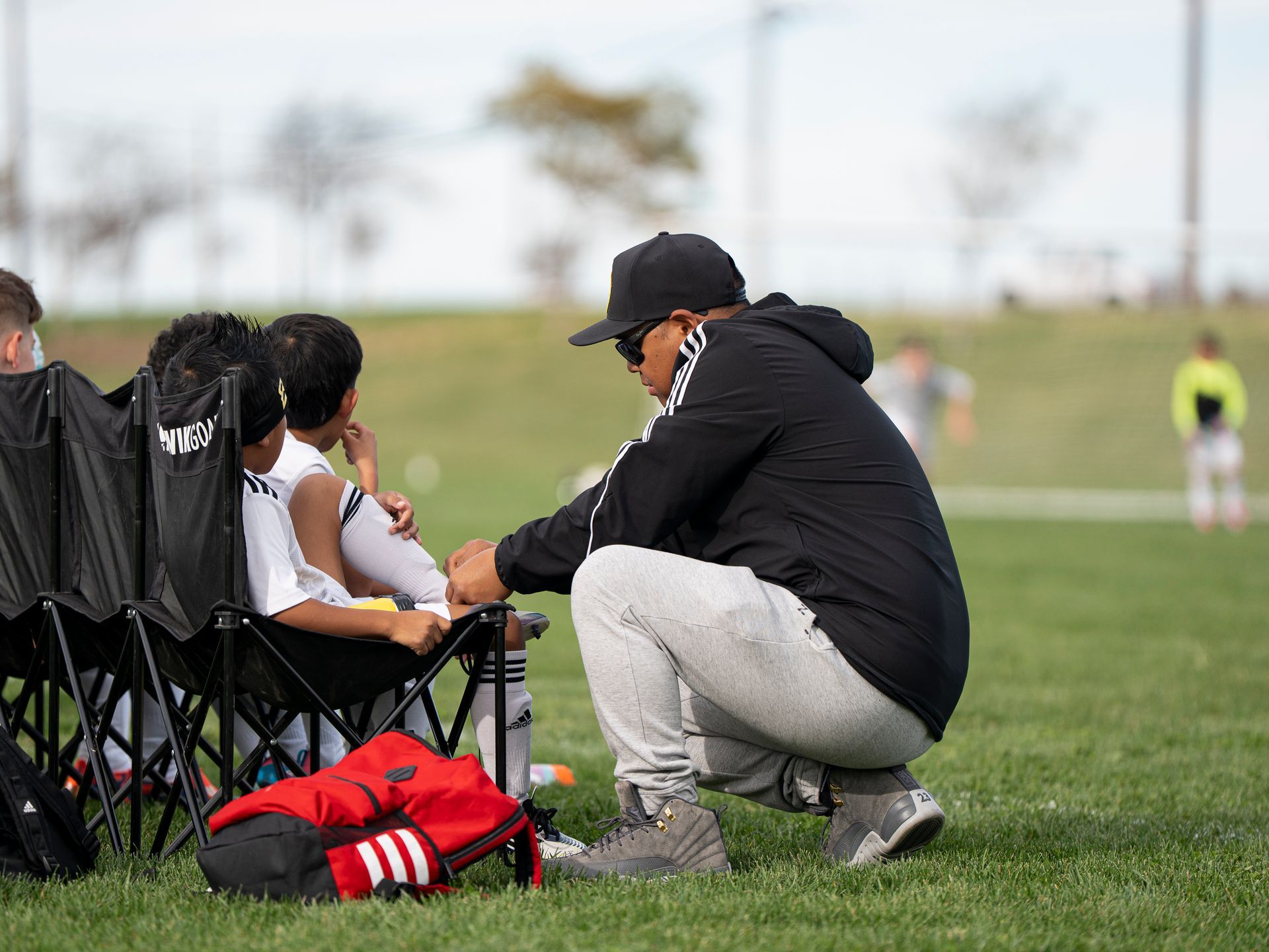 Soccer coach talking to players on sideline. Coach kneels, wearing a black jacket and cap. Players sit on folding chairs.