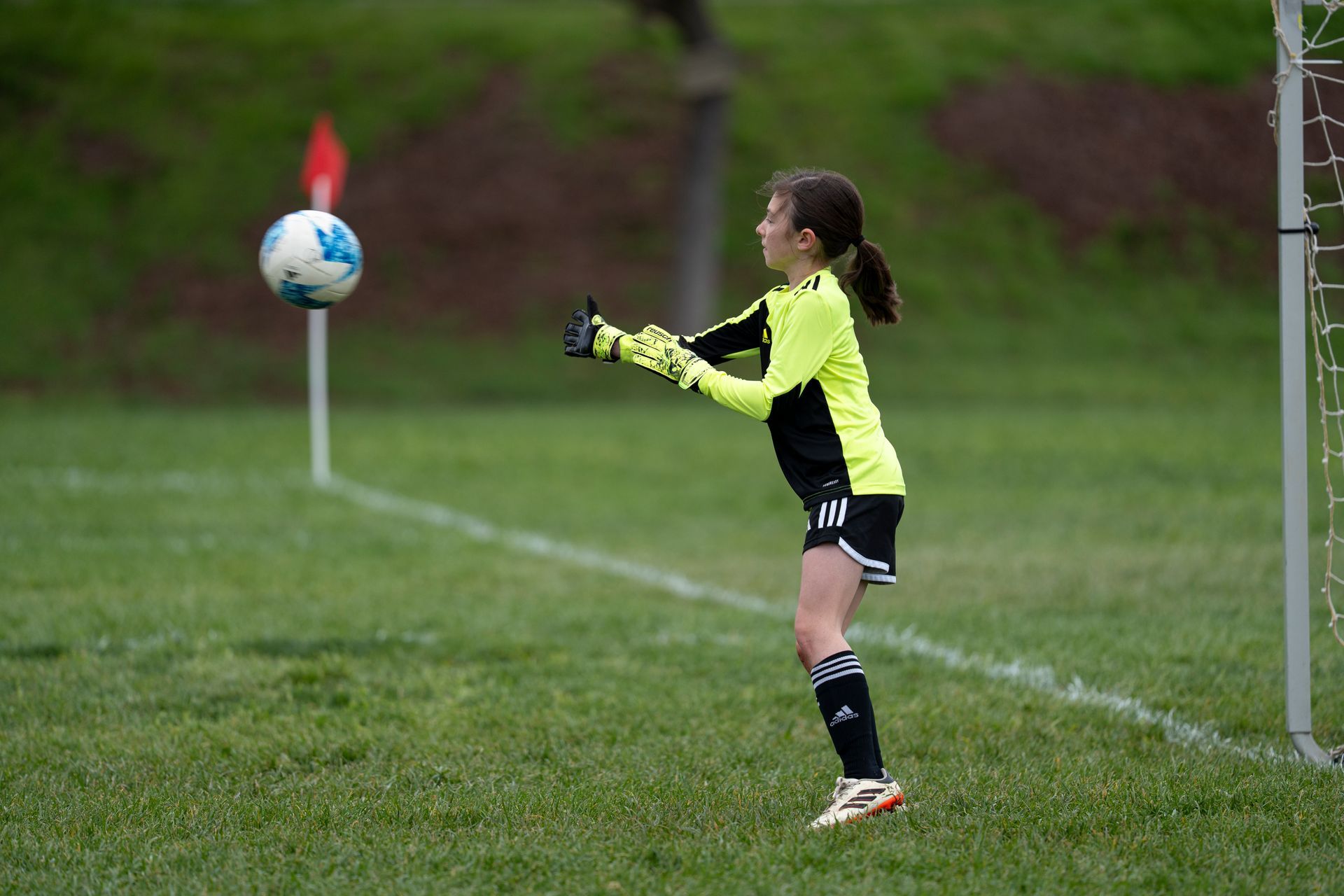Soccer goalie in neon yellow and black, reaching for the ball near a goal post on a green field.