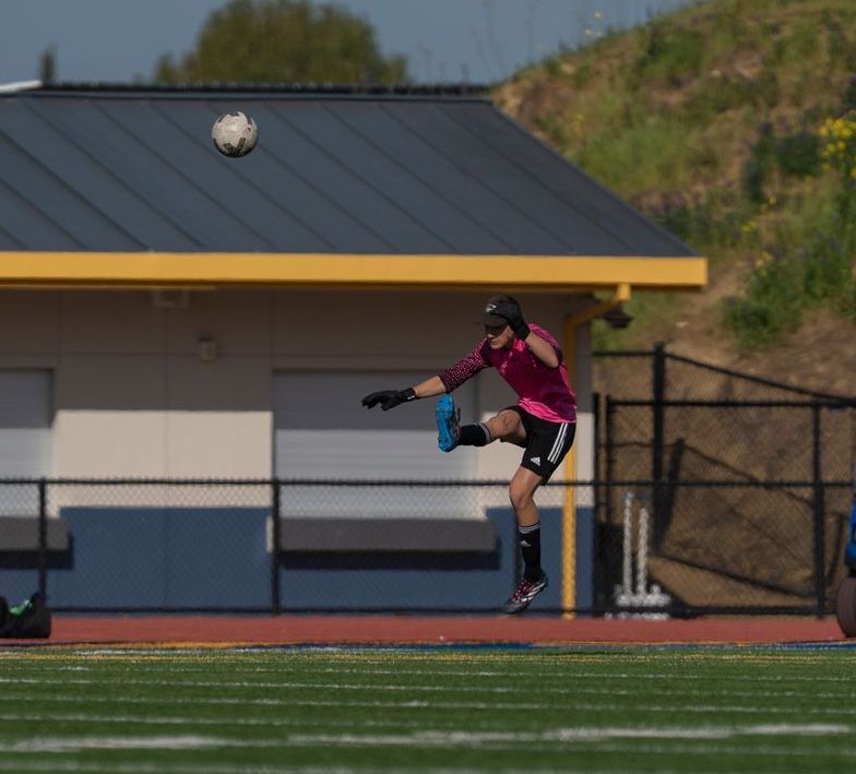 Soccer player in pink uniform kicks a soccer ball. Green field, building, and sky in background.