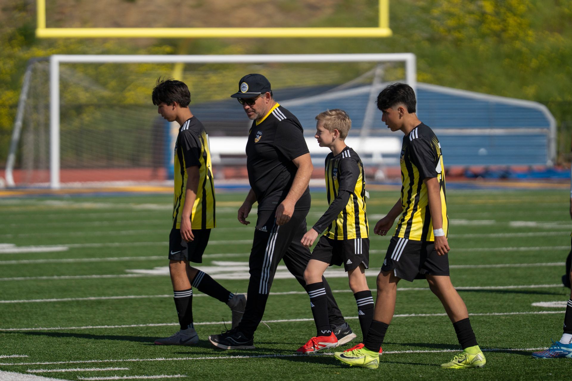 Soccer coach and players walking on field, wearing black and yellow uniforms.