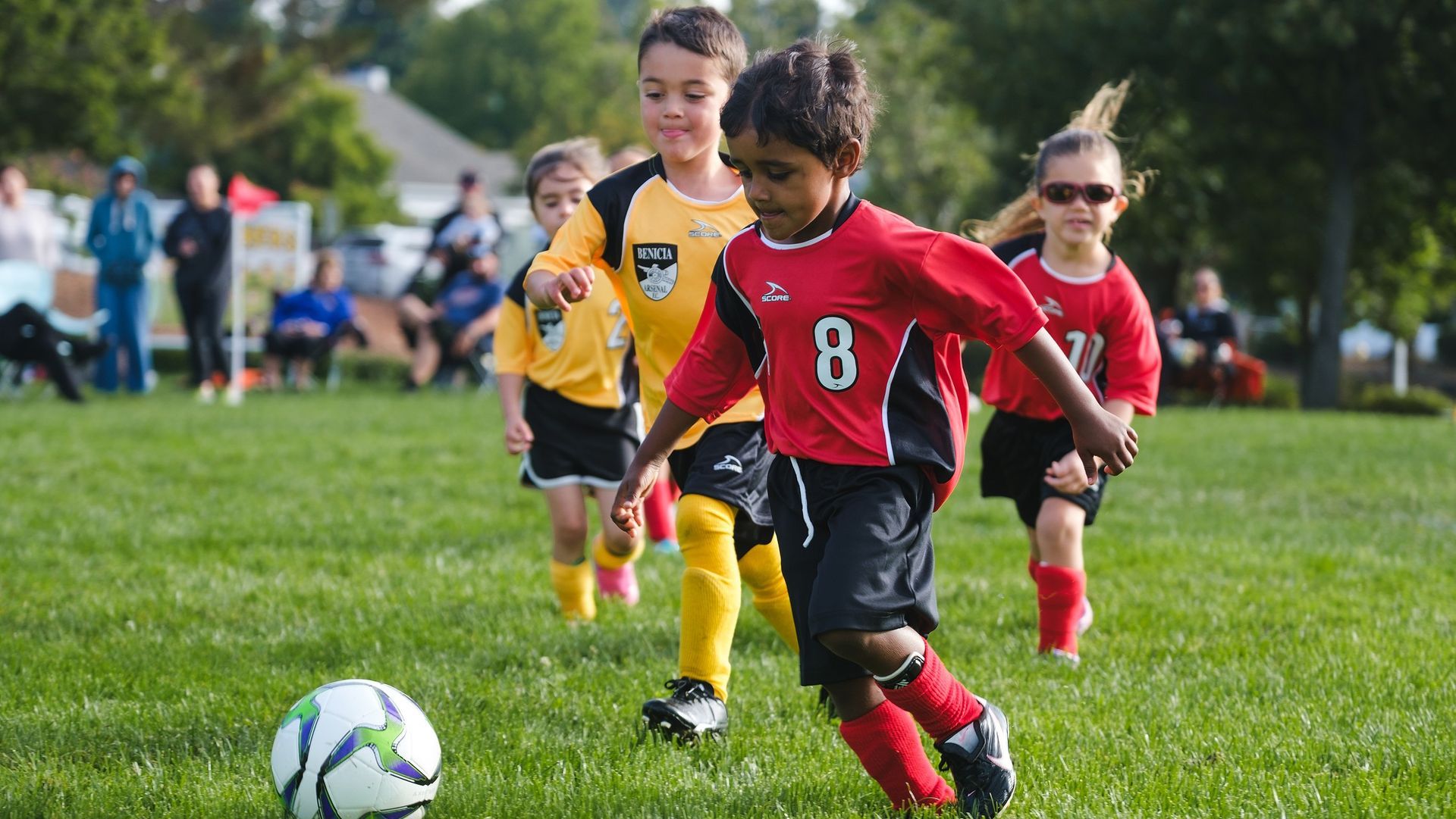 Kids playing soccer on a grassy field; one child in a red jersey dribbles the ball.