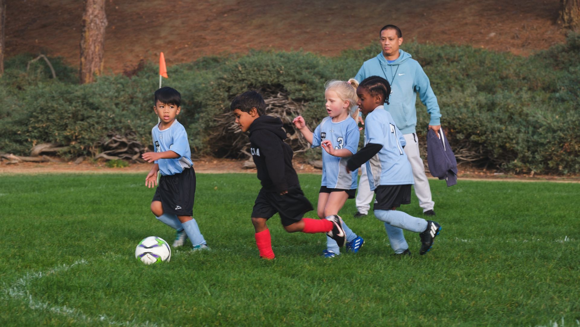 Children playing soccer on a grassy field, coach watches, one child kicks the ball.