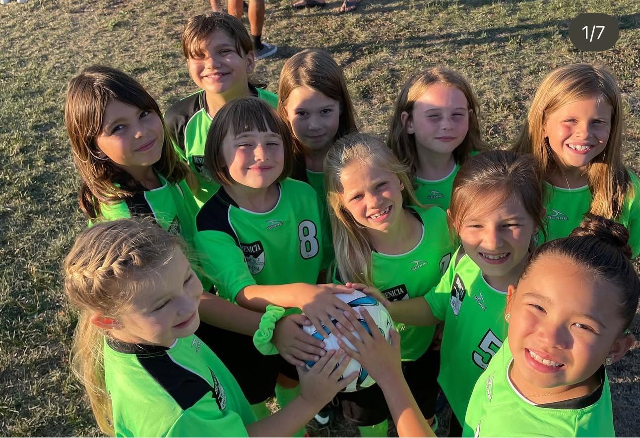 Soccer team in green jerseys huddling and smiling, holding a soccer ball on a field.