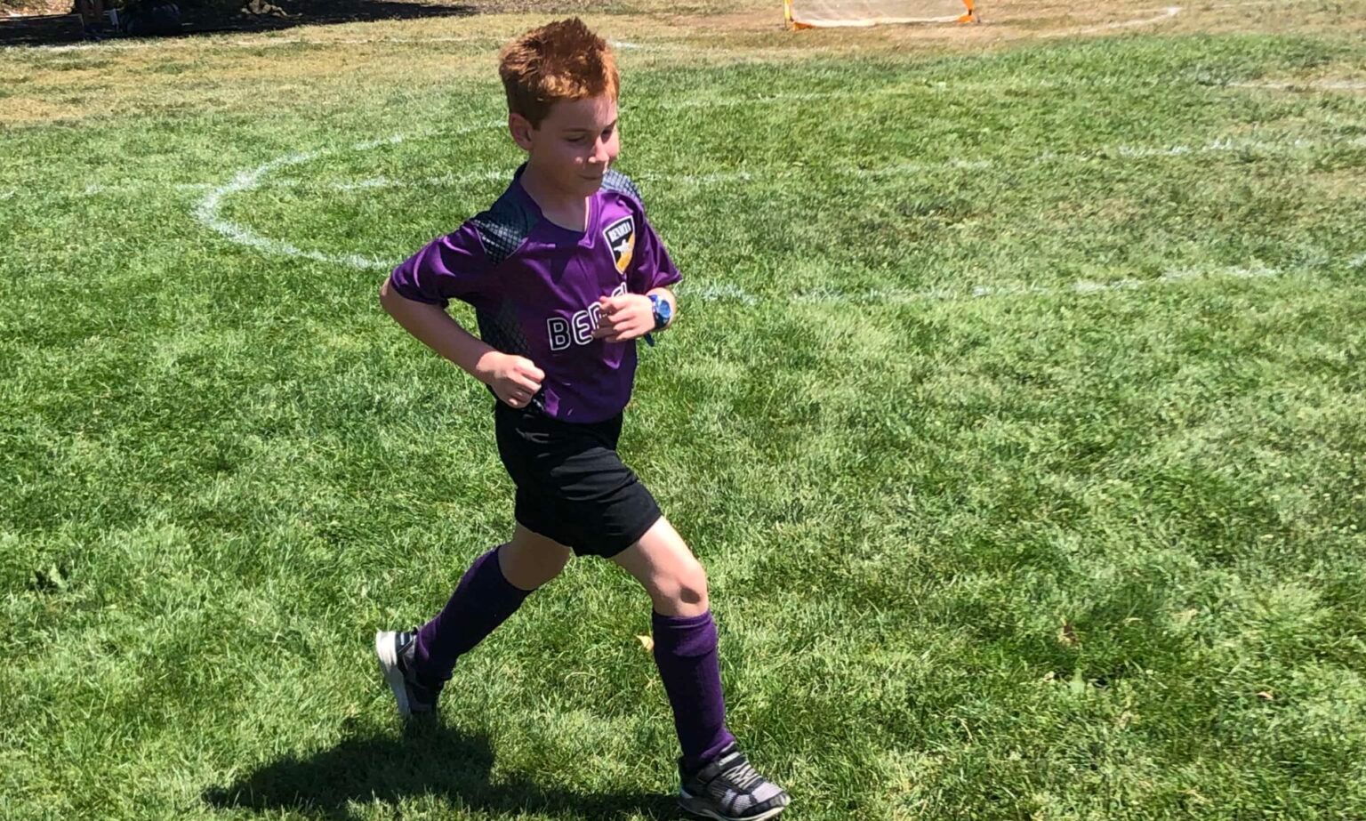 Boy in purple soccer uniform running on a grassy field.