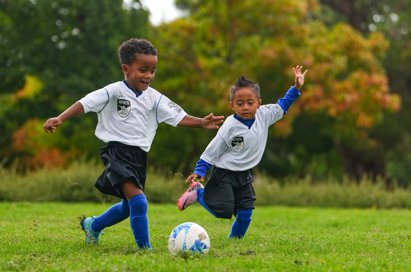 Two children in soccer uniforms playing soccer on a grassy field; one kicking the ball, other reaching.