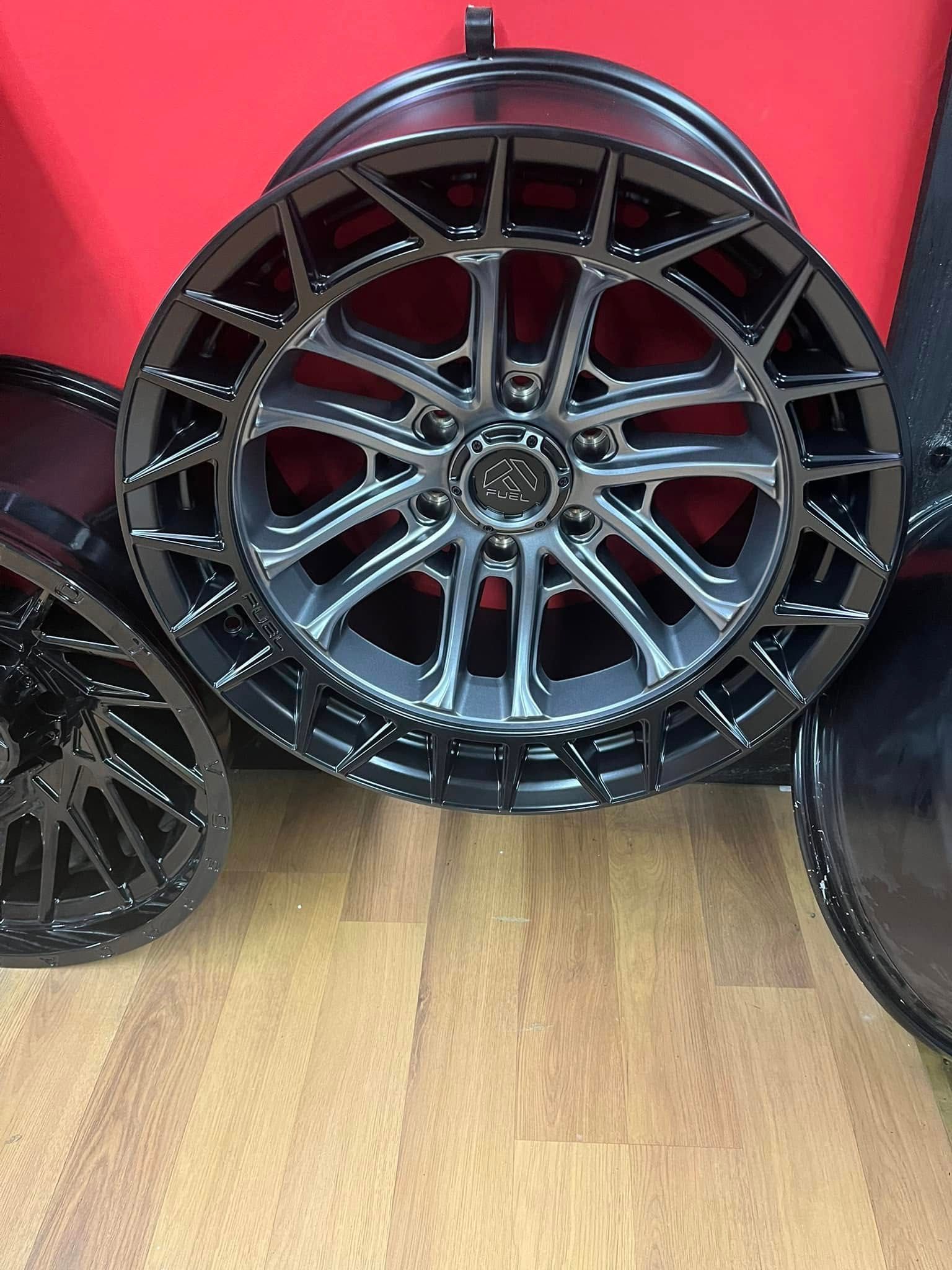 Black car rim with intricate geometric design, displayed indoors on wooden flooring against a red wall.