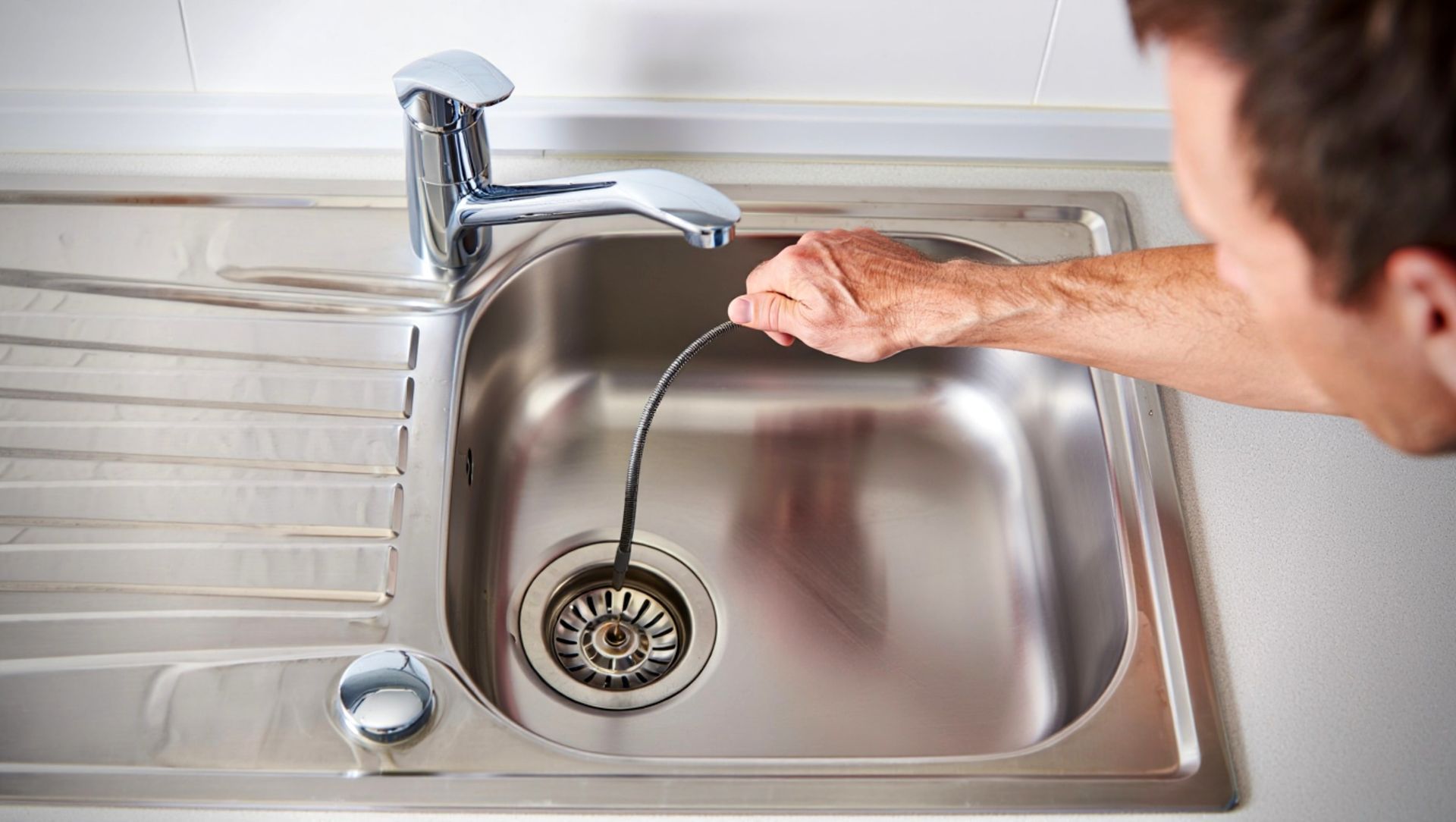Man unclogging a kitchen sink with a snake tool. The sink is stainless steel.