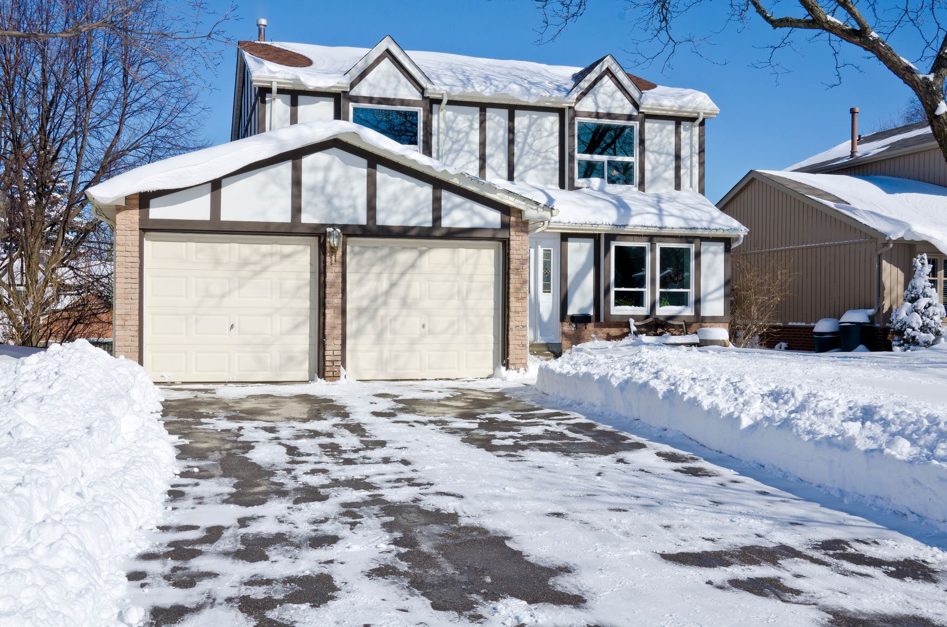 Snow-covered house with a two-car garage. Driveway partially cleared of snow, sunny day.