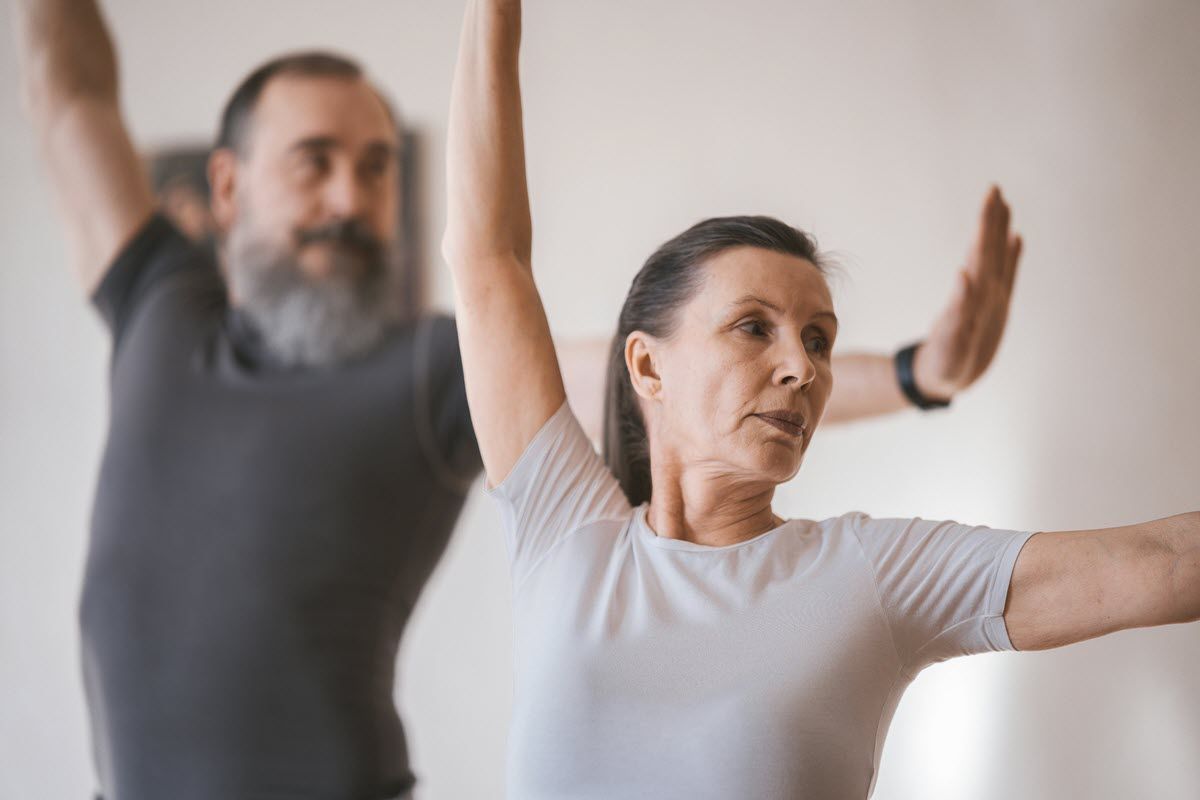Two people in a bright room performing a yoga pose with arms extended, focusing on their movements.
