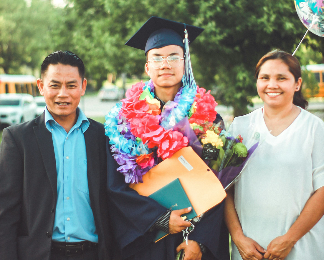 Graduate in cap and gown holding diploma, smiling with two people outdoors