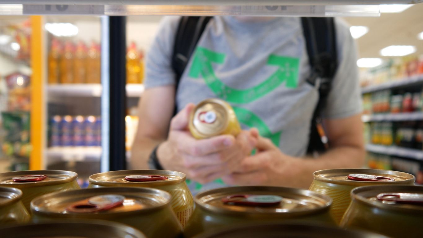 A person in a gray t-shirt examines a gold-colored can while shopping in a grocery store aisle filled with drinks.
