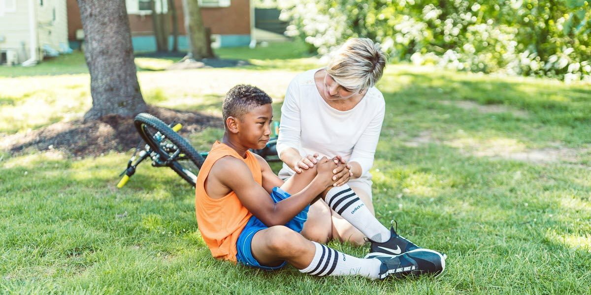 Woman helping boy with shin injury sit on grass near a bike in a sunny park