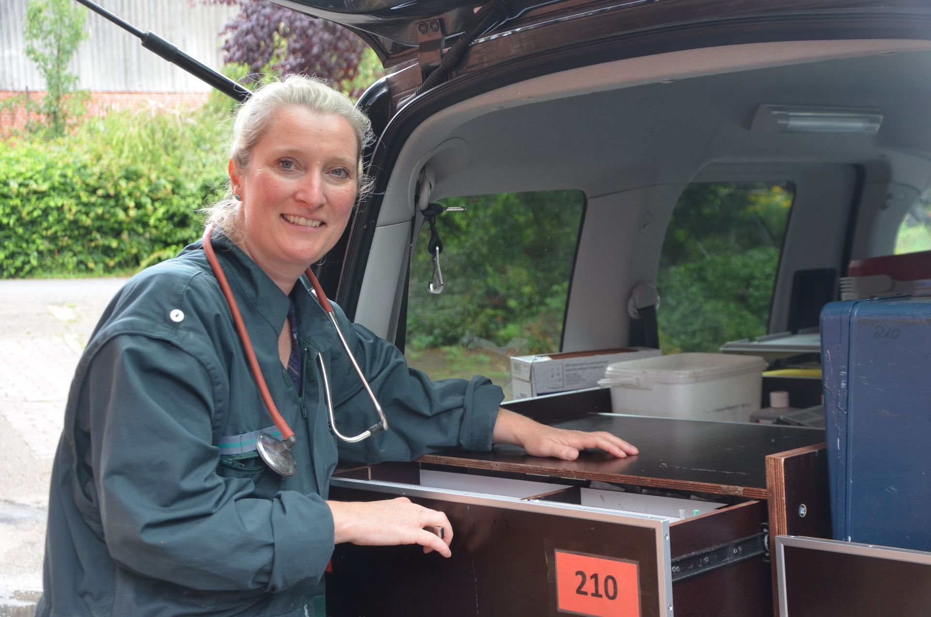 Tierarzt in grüner Uniform mit Stethoskop, lächelnd neben einem Fahrzeuginnenraum mit Ausrüstung.