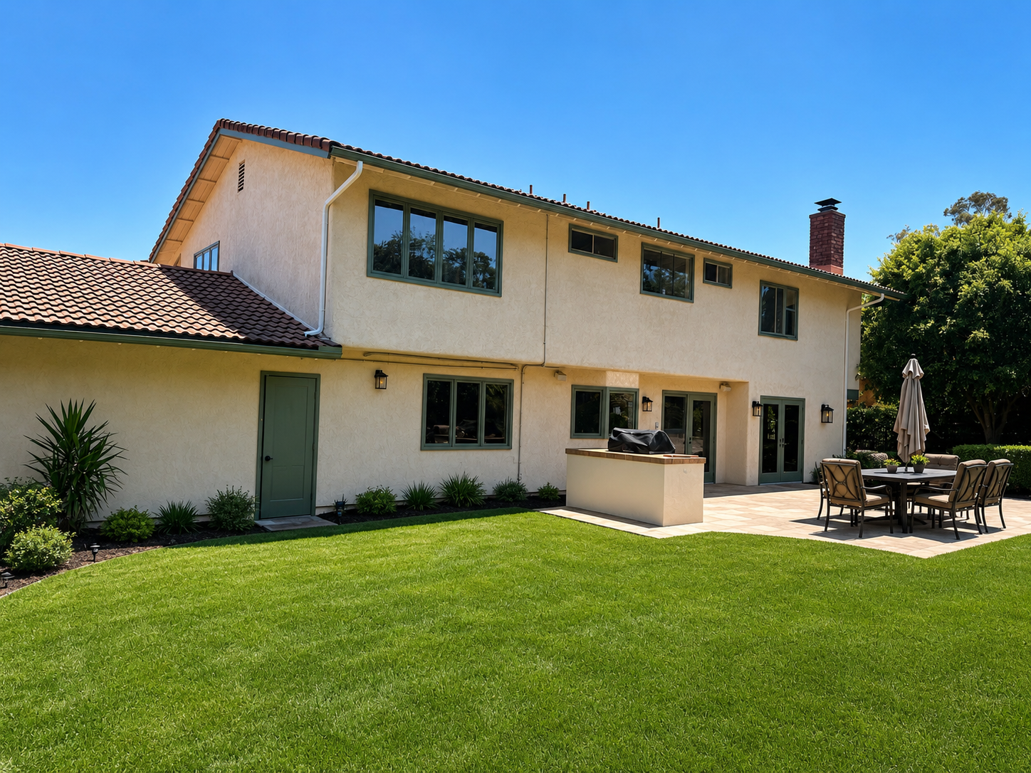 Two-story beige house with green lawn, patio table, and clear blue sky.