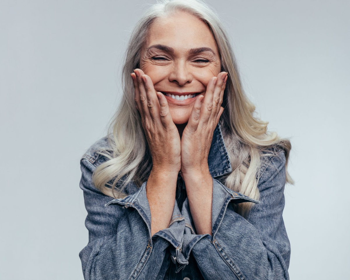 Smiling older woman with long gray hair, cupping her cheeks with both hands, wearing a denim jacket.