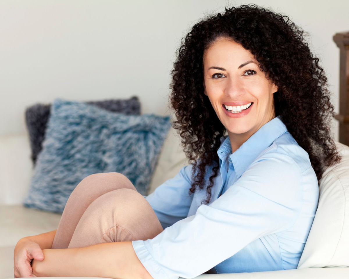 Woman with curly hair smiles, seated on a couch, wearing a light blue shirt and tan pants.