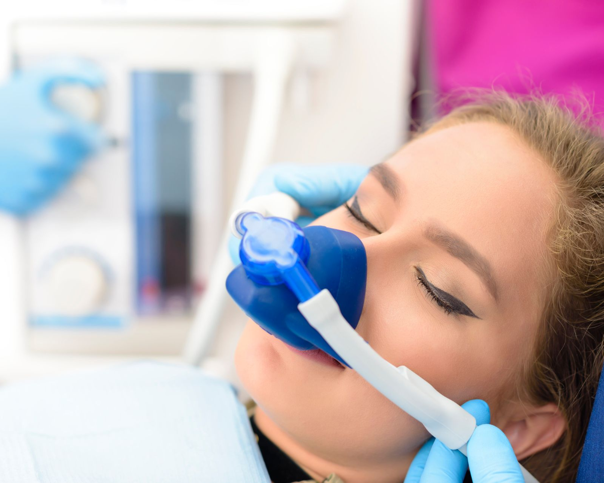 Woman receiving dental sedation; blue mask over nose, eyes closed, in a dental office.