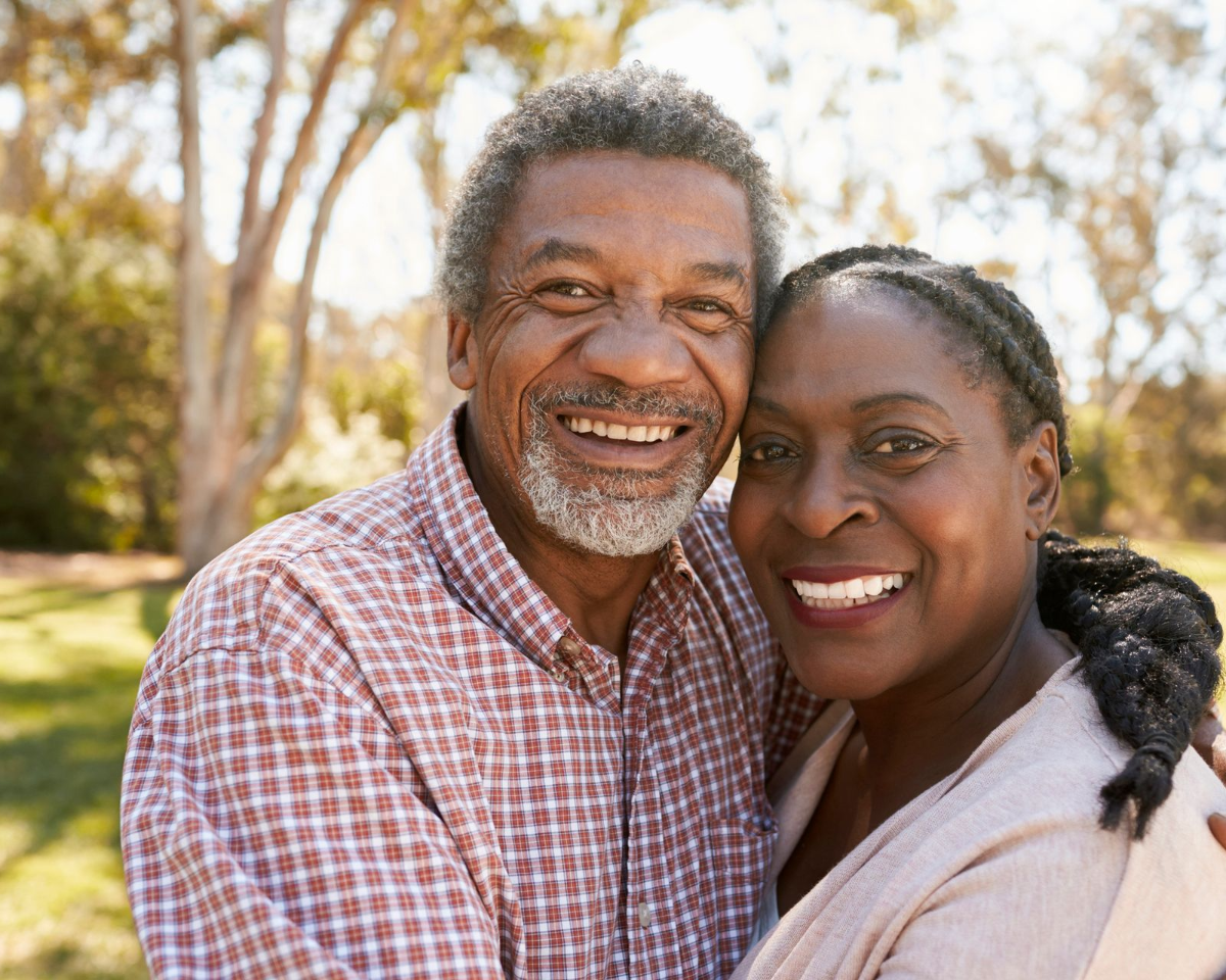 Smiling older African-American couple embracing outdoors in a park. The man wears a plaid shirt, the woman a light jacket.