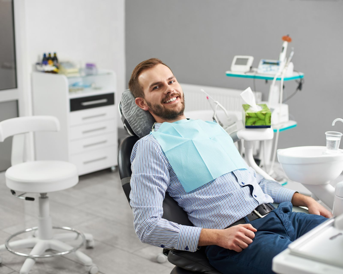 Smiling man in a dental chair wearing a bib, in a bright dentist's office.
