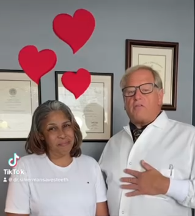A woman and a man stand together in front of a wall decorated with hearts. The man holds his hand to his chest.