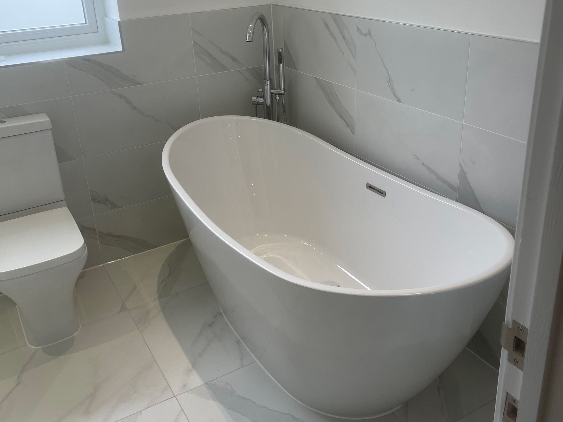 White freestanding bathtub and toilet in a modern bathroom with grey tile walls and floors.