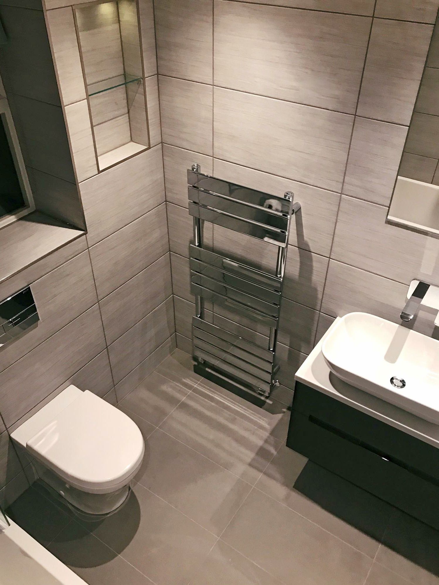 Modern gray tiled bathroom with toilet, sink, towel warmer, and recessed shelf.