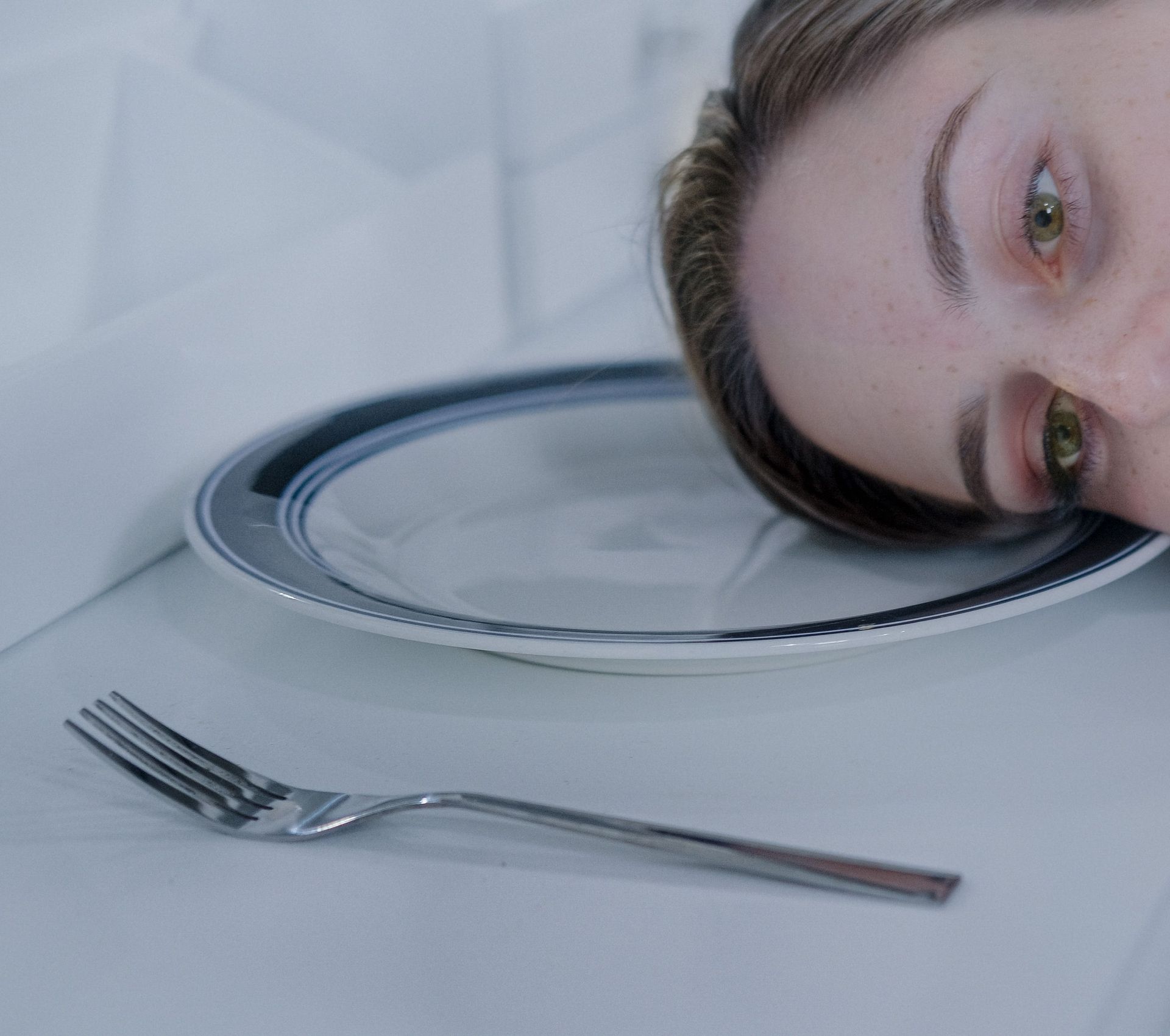A person’s face rests beside an empty plate and fork on a white table.